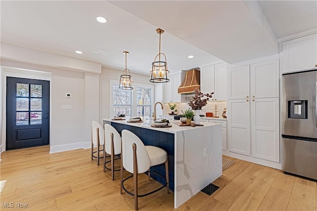 kitchen with a kitchen island with sink, white cabinets, hanging light fixtures, wall chimney exhaust hood, and stainless steel fridge with ice dispenser