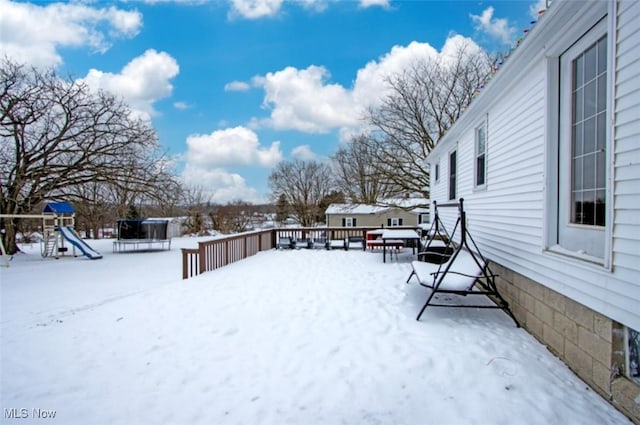 snowy yard with a trampoline, a playground, and a deck