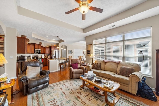 living room featuring a raised ceiling, ceiling fan, light hardwood / wood-style flooring, and a textured ceiling