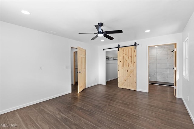 unfurnished bedroom featuring dark wood-type flooring, a closet, connected bathroom, a barn door, and ceiling fan