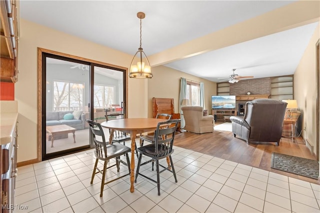 tiled dining area with ceiling fan with notable chandelier, a brick fireplace, and beam ceiling