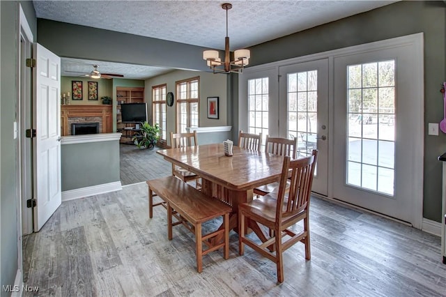 dining room featuring light wood-type flooring, a healthy amount of sunlight, a brick fireplace, a textured ceiling, and ceiling fan with notable chandelier