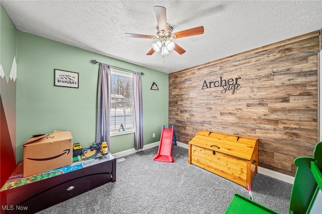 recreation room featuring a ceiling fan, a textured ceiling, carpet, wood walls, and baseboards