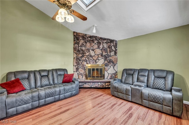 living room with ceiling fan, light wood-type flooring, a stone fireplace, and vaulted ceiling with skylight