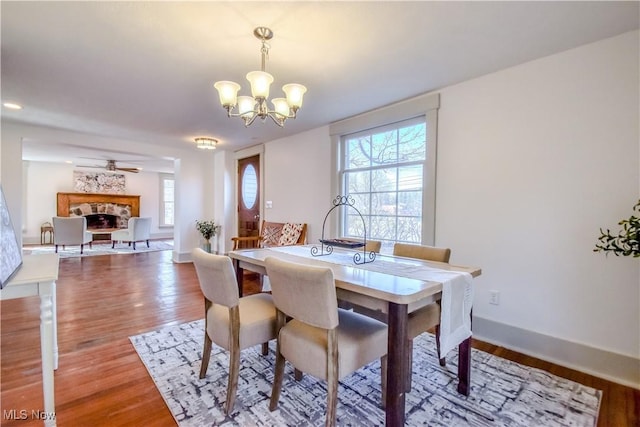 dining space featuring ceiling fan with notable chandelier, a fireplace, baseboards, and light wood-style floors