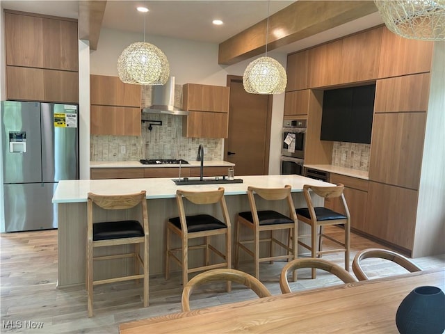 kitchen with stainless steel appliances, hanging light fixtures, wall chimney exhaust hood, light hardwood / wood-style flooring, and sink