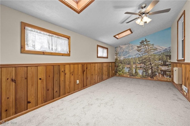 carpeted empty room featuring ceiling fan, radiator heating unit, lofted ceiling with skylight, and wooden walls