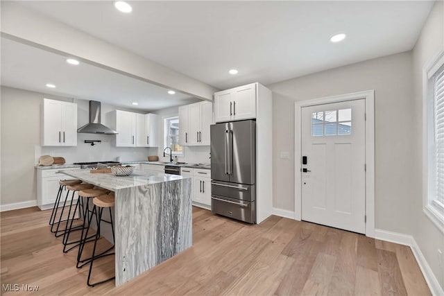 kitchen with white cabinetry, high end fridge, a kitchen island, wall chimney exhaust hood, and light stone counters