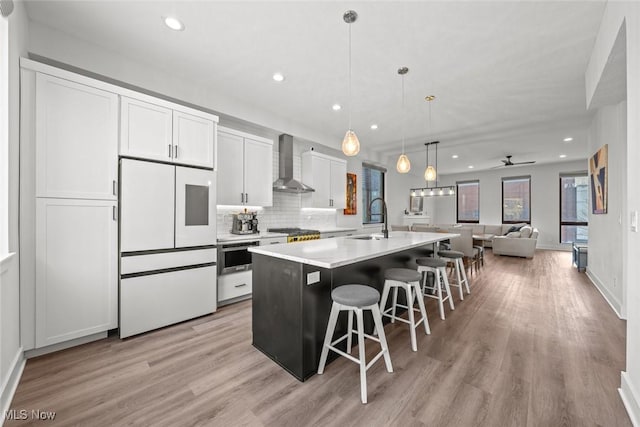 kitchen featuring appliances with stainless steel finishes, an island with sink, white cabinets, hanging light fixtures, and wall chimney exhaust hood