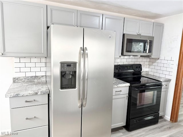 kitchen with light wood-type flooring, appliances with stainless steel finishes, gray cabinets, light stone countertops, and backsplash