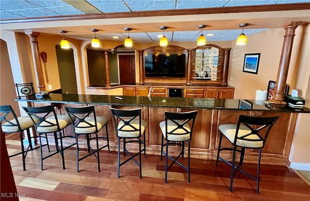 bar with decorative light fixtures, hardwood / wood-style floors, dark stone counters, and decorative columns