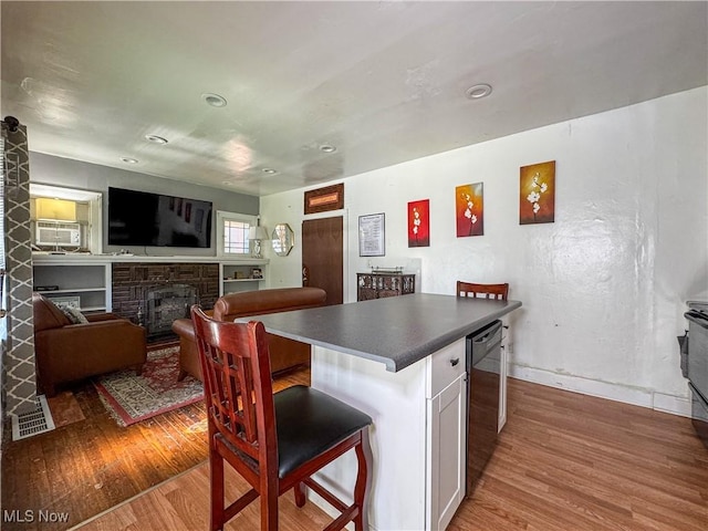 kitchen featuring a brick fireplace, a kitchen breakfast bar, dishwasher, wood-type flooring, and white cabinets