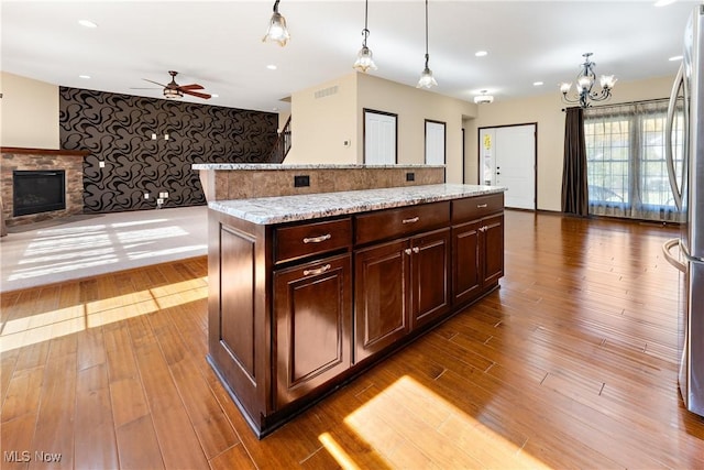 kitchen with a fireplace, a center island, dark brown cabinetry, hanging light fixtures, and light stone countertops