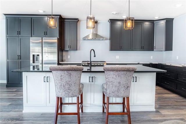 kitchen with hanging light fixtures, wall chimney range hood, dark hardwood / wood-style flooring, a center island with sink, and stainless steel fridge