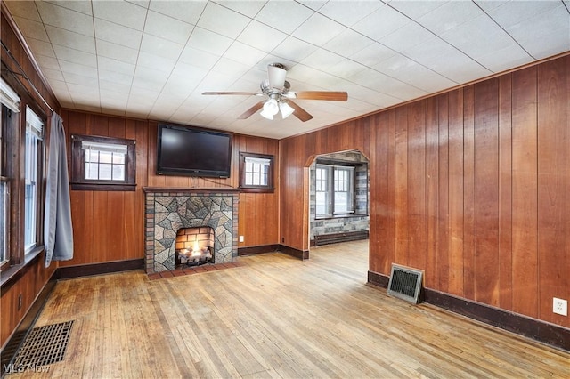 unfurnished living room featuring a stone fireplace, wooden walls, a baseboard radiator, light wood-type flooring, and ceiling fan