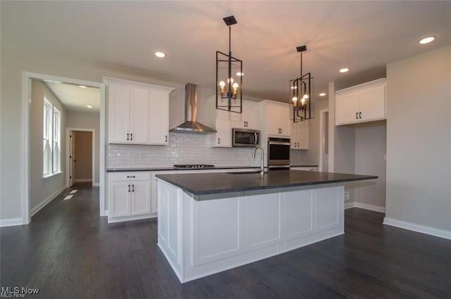 kitchen with hanging light fixtures, an island with sink, wall chimney range hood, and white cabinets