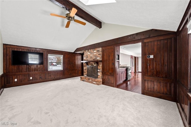 unfurnished living room with dark colored carpet, wood walls, a brick fireplace, ceiling fan, and vaulted ceiling with skylight