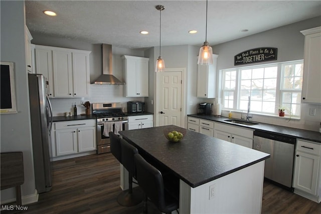 kitchen featuring a sink, stainless steel appliances, dark wood-type flooring, dark countertops, and wall chimney range hood