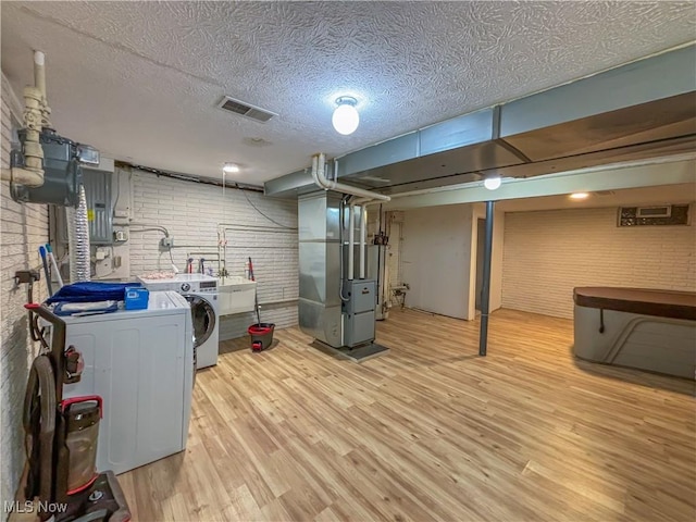 basement featuring washer and clothes dryer, heating unit, a textured ceiling, brick wall, and light wood-type flooring