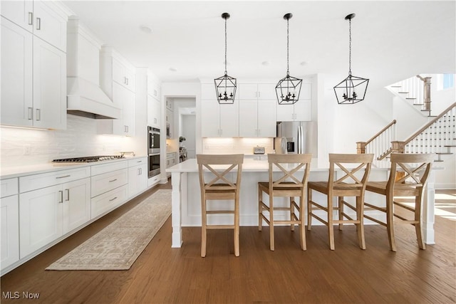 kitchen with appliances with stainless steel finishes, a kitchen island with sink, custom range hood, white cabinets, and a kitchen bar