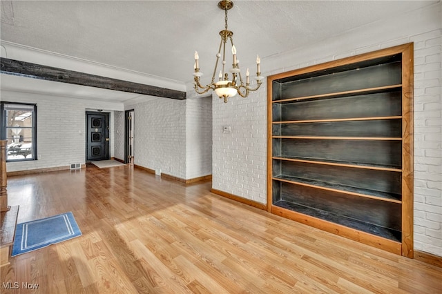 unfurnished dining area featuring brick wall, hardwood / wood-style floors, a chandelier, a textured ceiling, and beam ceiling