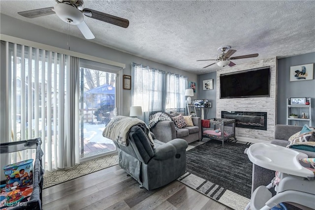 living room featuring ceiling fan, wood-type flooring, a stone fireplace, and a textured ceiling