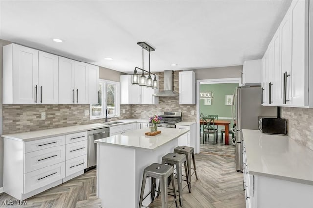 kitchen with wall chimney range hood, sink, appliances with stainless steel finishes, white cabinets, and a kitchen island