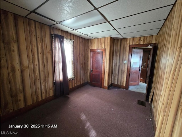 empty room featuring a paneled ceiling, dark colored carpet, and wood walls