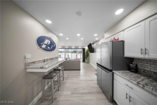 kitchen featuring white cabinetry, light stone counters, stainless steel fridge, a kitchen breakfast bar, and backsplash