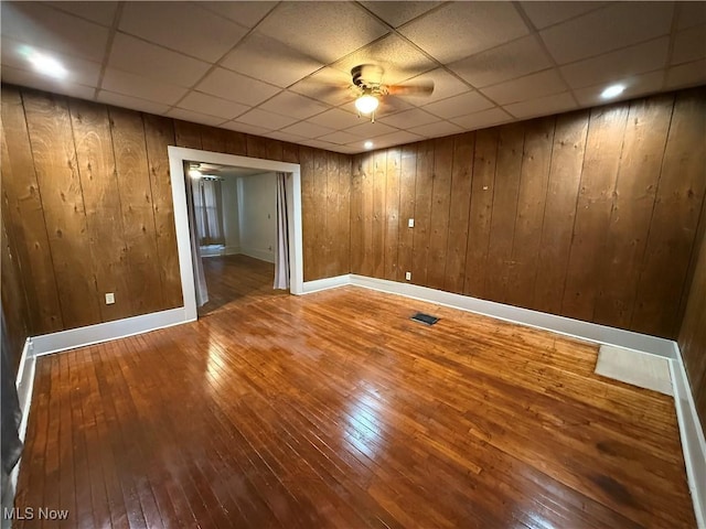 empty room featuring ceiling fan, a paneled ceiling, wood-type flooring, and wooden walls