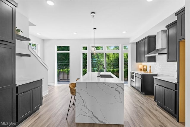 kitchen featuring wall chimney exhaust hood, sink, light wood-type flooring, a kitchen island with sink, and decorative backsplash