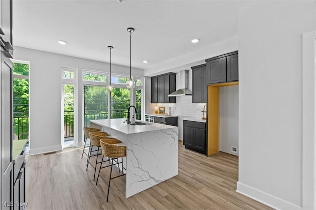 kitchen with sink, pendant lighting, a kitchen island with sink, decorative backsplash, and wall chimney range hood