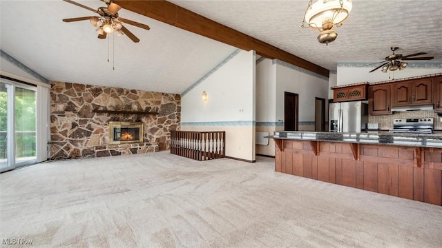 kitchen featuring appliances with stainless steel finishes, vaulted ceiling with beams, a kitchen breakfast bar, a stone fireplace, and light colored carpet