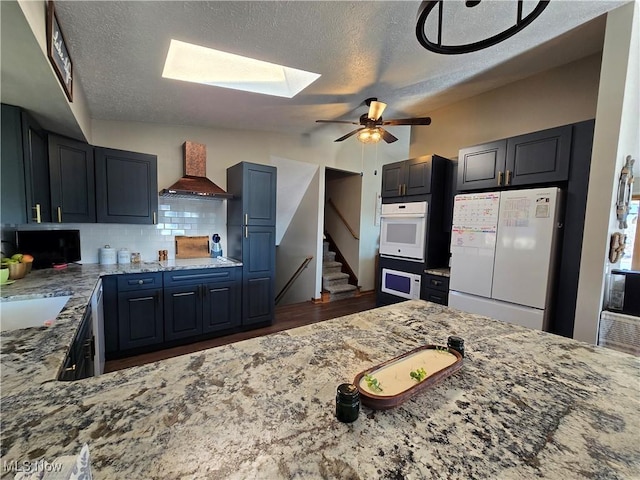 kitchen featuring white appliances, backsplash, lofted ceiling with skylight, light stone counters, and custom exhaust hood