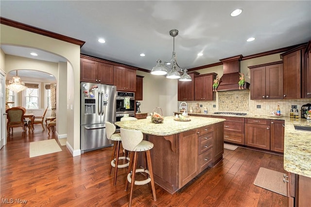 kitchen with hanging light fixtures, stainless steel appliances, light stone counters, custom range hood, and an island with sink