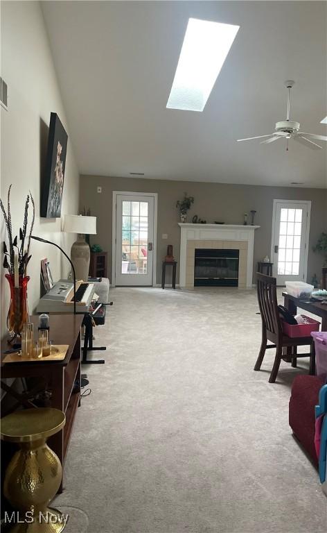 carpeted home office featuring a tile fireplace, a ceiling fan, visible vents, and vaulted ceiling with skylight