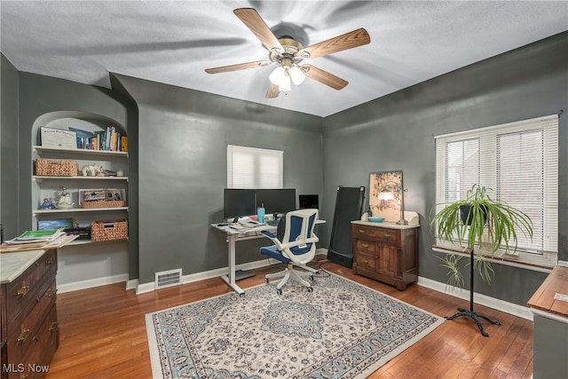 office area with wood-type flooring, a textured ceiling, ceiling fan, and built in shelves