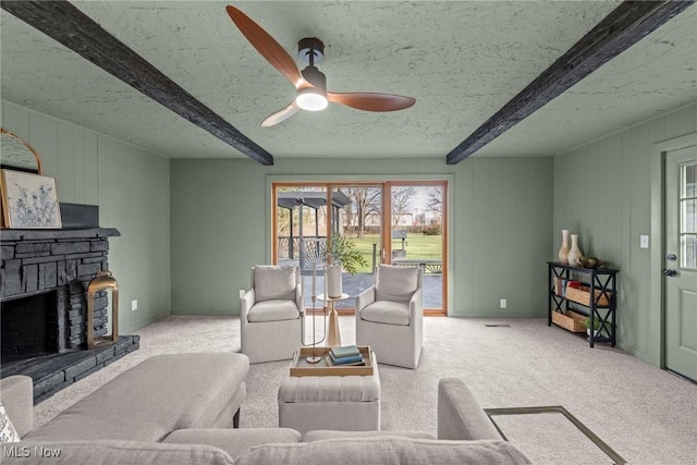 carpeted living room featuring beamed ceiling, ceiling fan, a stone fireplace, and a textured ceiling