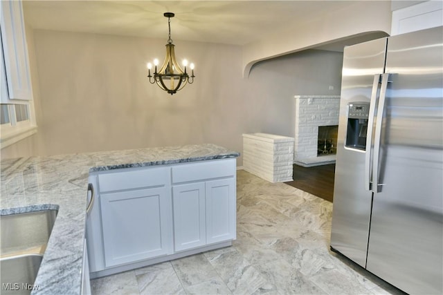 kitchen with stainless steel refrigerator with ice dispenser, hanging light fixtures, a fireplace, light stone countertops, and white cabinets