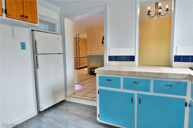 kitchen with blue cabinets, hanging light fixtures, white fridge, tile counters, and a notable chandelier