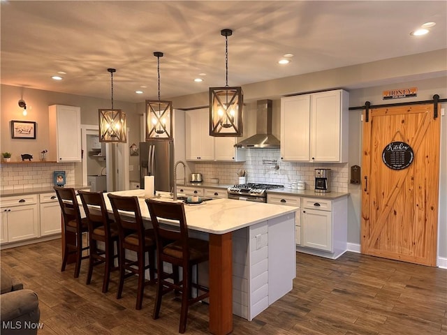 kitchen featuring a center island with sink, wall chimney range hood, stainless steel appliances, a barn door, and white cabinets