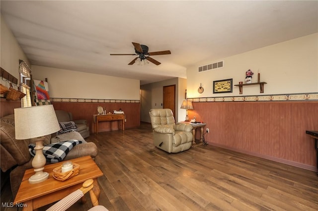 living room with a wainscoted wall, a ceiling fan, visible vents, and dark wood-type flooring