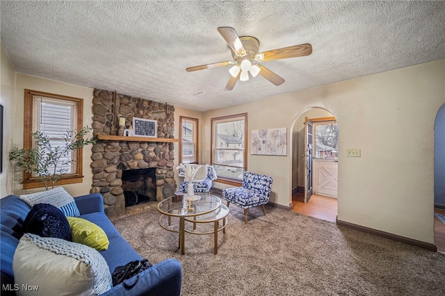 living room featuring ceiling fan, a stone fireplace, a textured ceiling, and carpet flooring