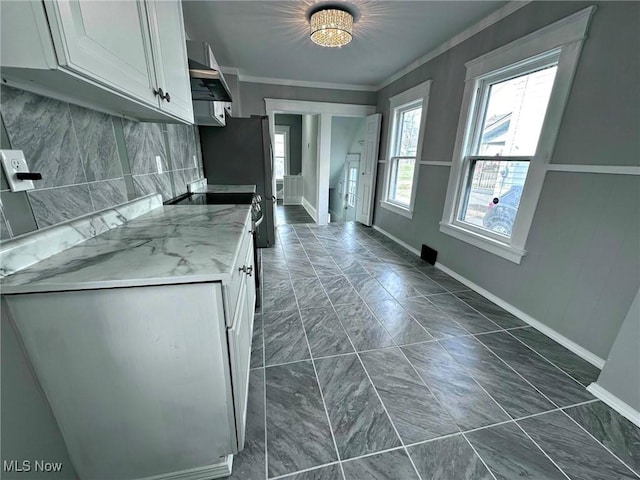 kitchen with wall chimney range hood, backsplash, ornamental molding, light stone countertops, and white cabinets