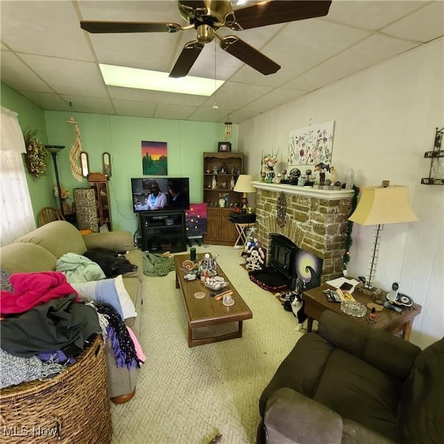 living room with a drop ceiling, a stone fireplace, and ceiling fan