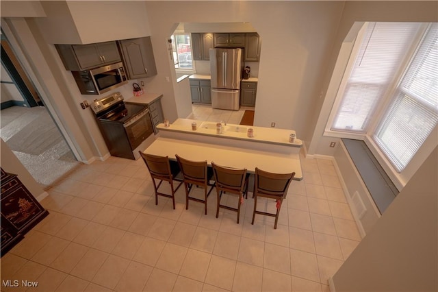 kitchen with light tile patterned floors, gray cabinets, stainless steel appliances, a kitchen breakfast bar, and kitchen peninsula