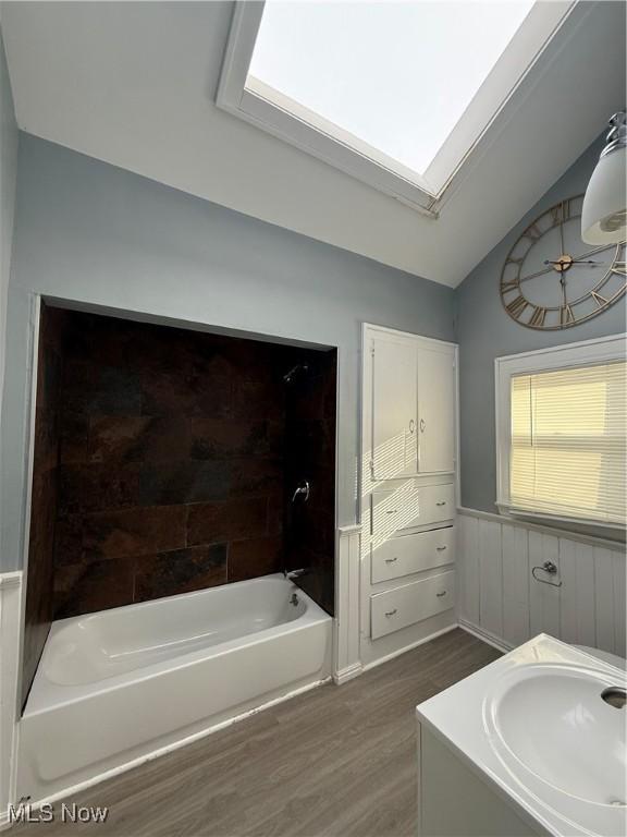 full bath featuring a tub to relax in, wainscoting, lofted ceiling with skylight, wood finished floors, and a sink