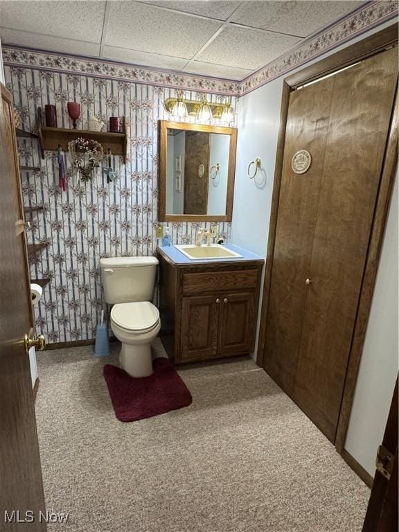 half bathroom featuring wallpapered walls, baseboards, a drop ceiling, toilet, and vanity