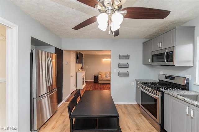 kitchen with gray cabinetry, a textured ceiling, appliances with stainless steel finishes, light stone countertops, and light hardwood / wood-style floors