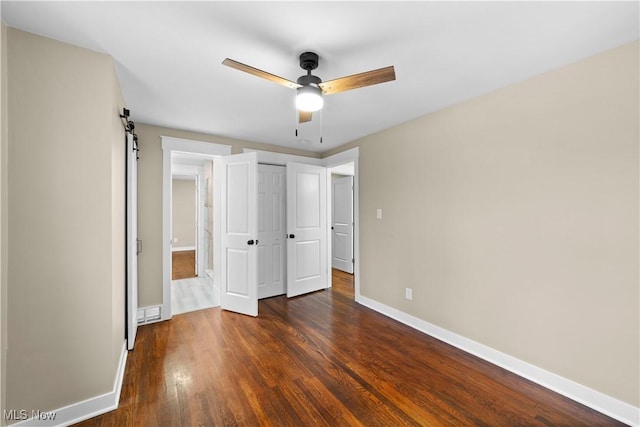 unfurnished bedroom featuring ceiling fan, a barn door, dark hardwood / wood-style flooring, and a closet